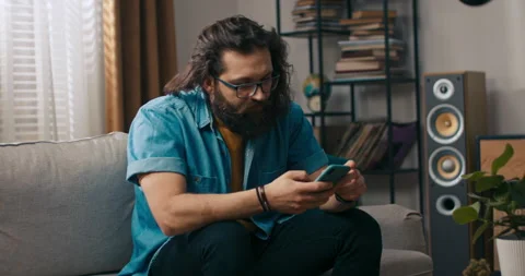 A bearded handsome guy is sitting on the couch with his dog in the living room 库存影片 218180060