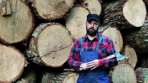 A bearded, handsome logger in a checked shirt stands with an axe at the sawmill Stock Footage 133429309