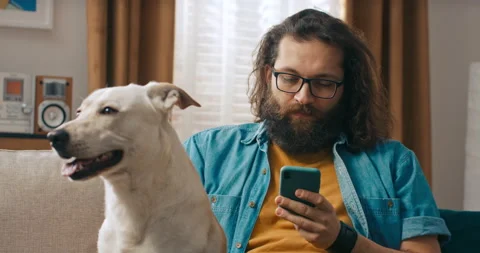 A bearded handsome man sits on the couch with his dog in the living room, which 스톡 동영상 218187094