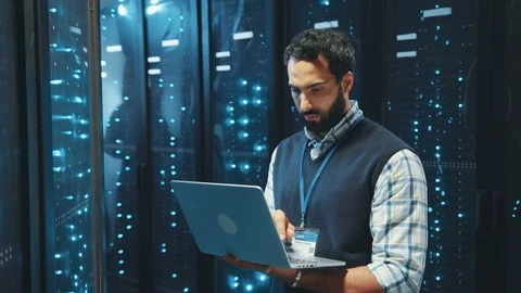 Bearded IT engineer typing on laptop running diagnostics of server rack Видео 119278851