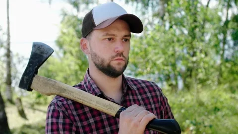 A bearded lumberjack in a cap with a big axe on his shoulder in the forest Stock Footage 147995693