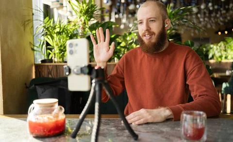 Bearded male influencer broadcasting using smartphone and tripod, sitting i.. Stock Photos