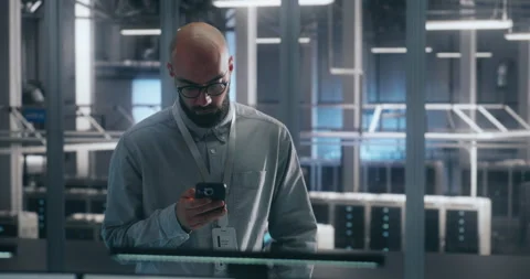 Bearded Male IT Engineer in Glasses Stands and Uses Smartphone to Check Critical Stock Footage 325426405