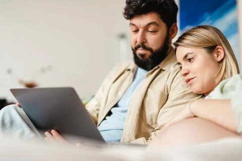 Bearded man and pregnant woman using laptop while sitting on couch Stock Photos