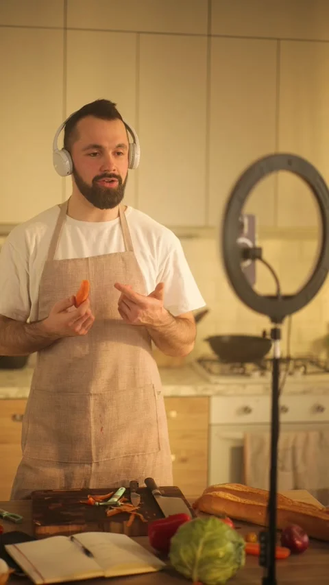 A bearded man in an apron records a cooking vlog in his kitchen. He speaks to Stock Footage 304611085