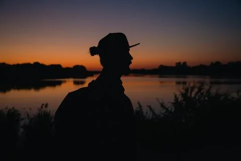 A bearded man with backpack and with cap at sunset near the lake. Stock Photos