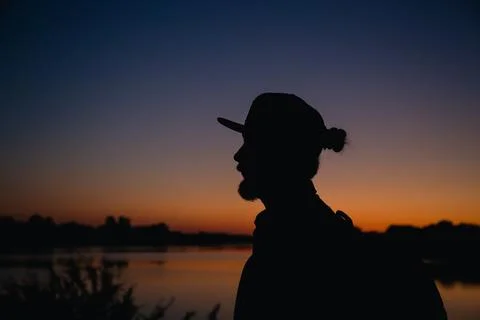 A bearded man with backpack and with cap at sunset near the lake. Stock Photos