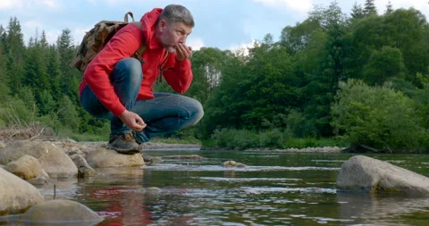 A bearded man with a backpack drinks water from a mountain river Stock Footage 157183902