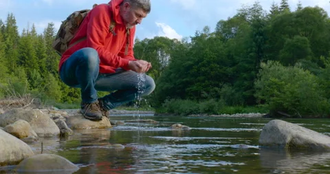 A bearded man with a backpack drinks water from a mountain river Stock Footage 157209119