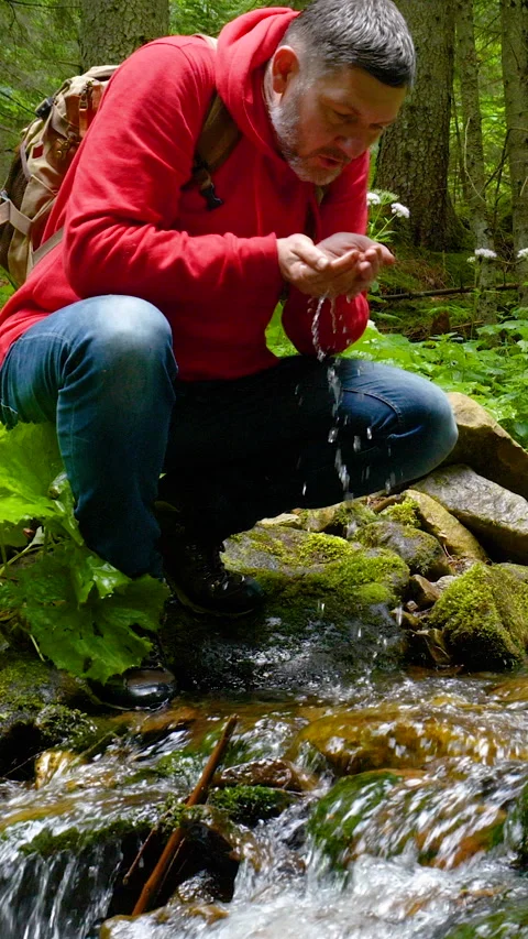 A bearded man with a backpack drinks water from a mountain stream 库存影片 334387650