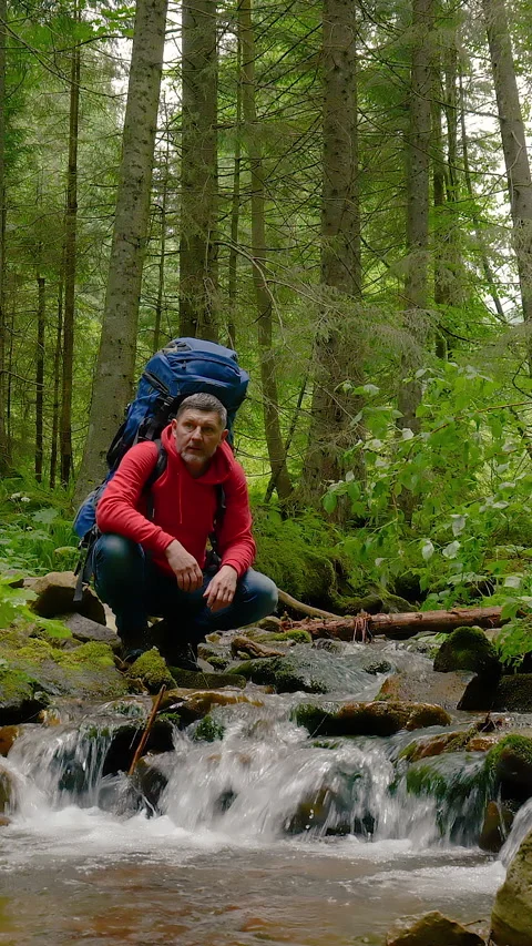 A bearded man with a backpack drinks water from a Mountain stream Video stock 336045028