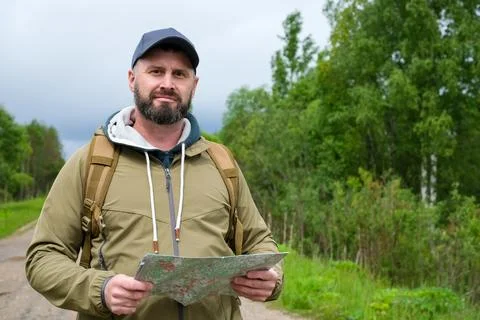 Bearded man with backpack holding map on rural road near forest and looking a Stock Photos