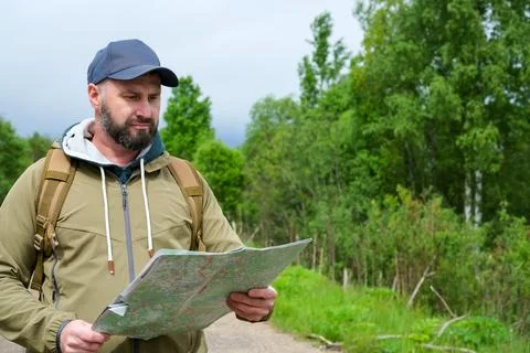 Bearded man with backpack reading paper map on rural forest road under cloudy 写真素材