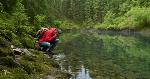 A bearded man with a backpack washes himself with water from a clear lake Stock Footage 156965799