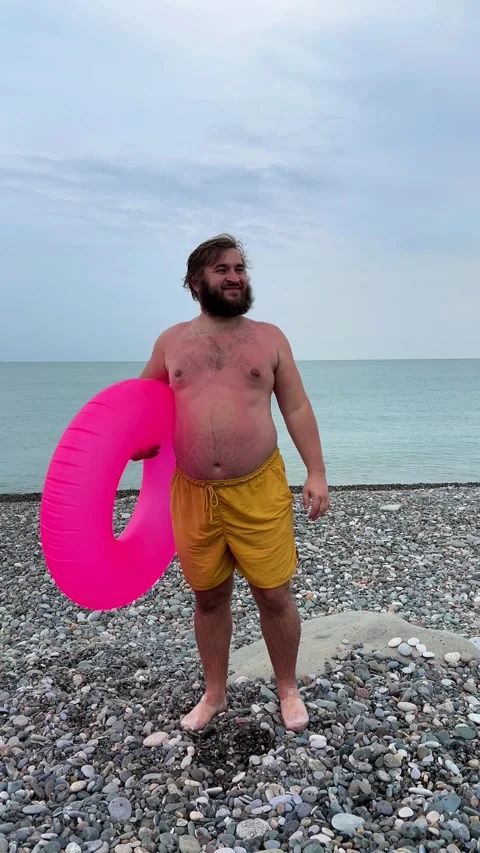 Bearded man with big belly with pink inflatable swim ring on pebble beach Stock Footage 281127532
