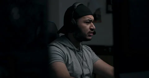 Bearded man in black cap sits on chair at computer desk in dark room. Guy in Stock Footage 214191394