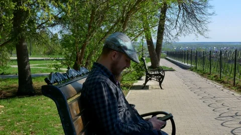 A bearded man in a cap sits in a park on a bench uses a phone sends a message Stock Footage 125959971