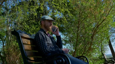 A bearded man in a cap sits in a park on a bench talking on the phone Stock Footage 125960089