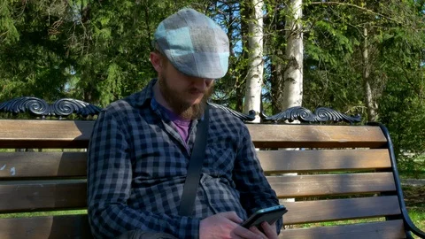 A bearded man in a cap sits in a park on a bench uses a phone sends a message Stock Footage 125960652
