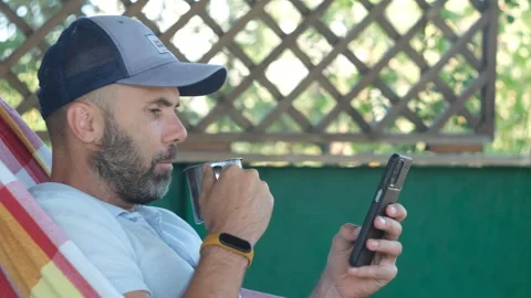 A bearded man in cap smartphone and metal cup relaxing in hammock on the terrass Stock Footage 252280632