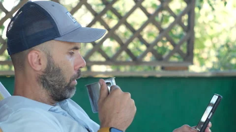 A bearded man in cap smartphone and metal cup relaxing in hammock on the terrass Stock Footage 252280927
