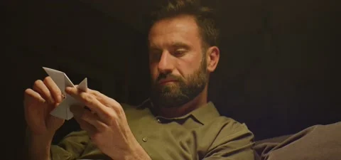 Bearded man on couch thinking, folding a bird from paper, evening Video stock 201143622