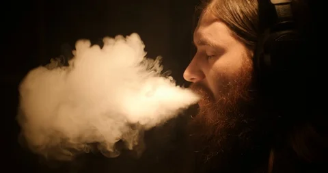 A bearded man in the darkness smokes an electronic cigarette and listens to Stock Footage 88057669