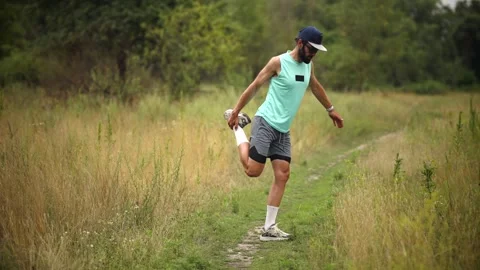 Bearded man doing standing quadriceps stretch on grassy trail in rural meadow Stock Footage 313951458