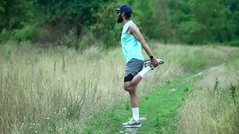 Bearded man doing standing quadriceps stretch on grassy trail during warm-up Stock Footage 313951466