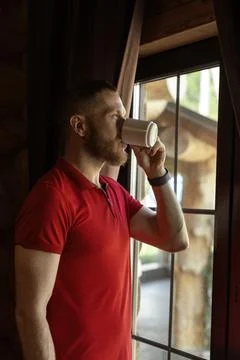 Bearded man drinking coffee from white mug by window wearing red polo shirt Stock Photos