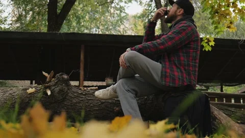 Bearded man drinking tea from a bowl sitting on a wooden stump in autumn park Stock Footage 220415406