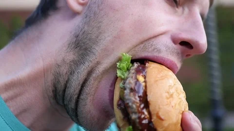 Bearded man eats with appetite a delicious and juicy burger in the garden. SLOW Stock Footage 77711629