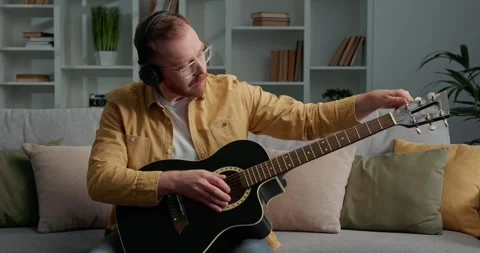 Bearded man with eyeglasses sitting on sofa, learning to play acoustic guitar. Stock Footage 249465728