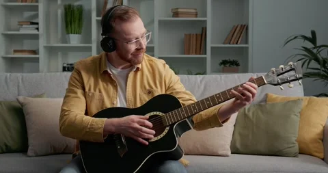 Bearded man with eyeglasses sitting on sofa, learning to play acoustic guitar. Stock Footage 249465764