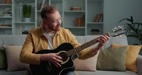 Bearded man with eyeglasses sitting on sofa, learning to play acoustic guitar. Stock Footage 249465863