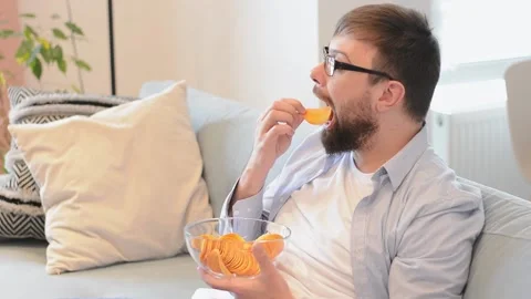 Bearded man in glasses eating chips watching football at home on the couch Stock Footage 200138991