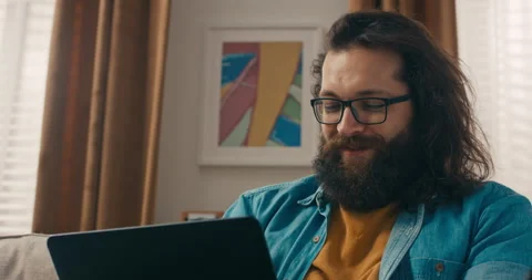 A bearded man with glasses sits on a couch with a laptop, working remotely Stock Footage 218211356