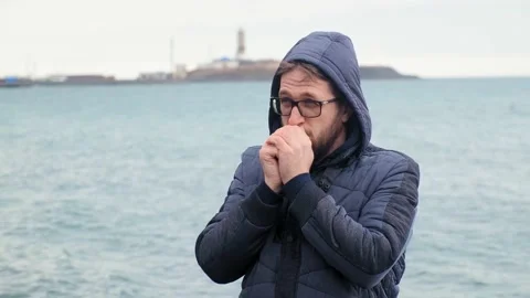 Bearded man in glasses warms hands standing against blue sea 库存影片 134268539