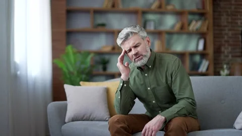 Bearded man with gray hair sitting on sofa at home suffering from headache.  Stock Footage 328706125