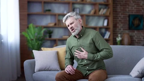 Bearded man with gray hair sitting on sofa at home suffering from chest pain  Stock Footage 328706147