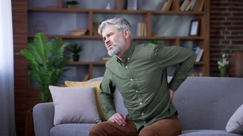 Bearded man with gray hair sitting on sofa at home suffering from back pain.  Stock Footage 328706304