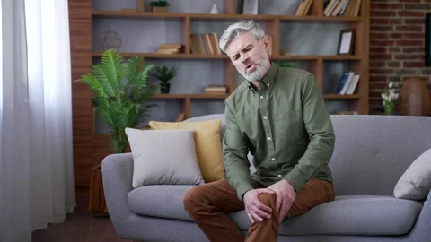 Bearded man with gray hair sitting on sofa at home suffering from knee pain.  Video stock 328706361
