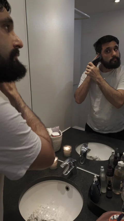 Bearded man grooming beard with electric trimmer in bathroom mirror Video stock 335637552