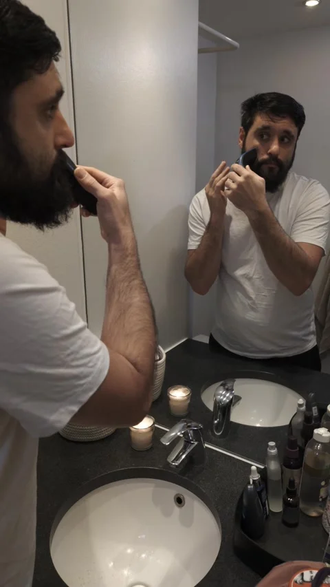 Bearded man grooming beard with electric trimmer in bathroom mirror Video stock 335638169