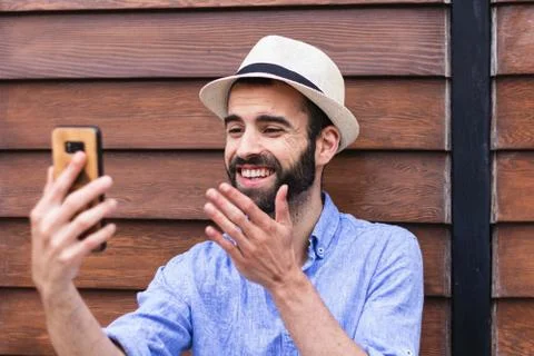 Bearded man with hat making a video call in front of a wooden wall Foto stock