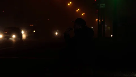 Bearded man in jacket and cap sits on the side of the road on the sidewalk and Stock Footage 120419527