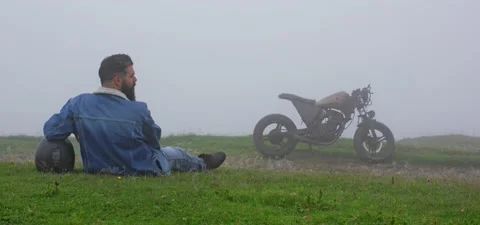 Bearded man in jeans on grass with old cafe racer motorcycle in mountains mist Stock Footage 140623550