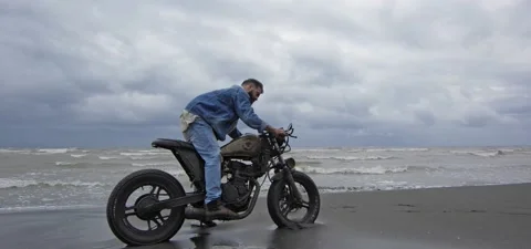 Bearded man in jeans on his old cafe racer motorcycle near sea, kickstarting Stock Footage 140624847