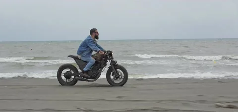 Bearded man in jeans rides old cafe racer motorcycle on sandy beach near sea Stock Footage 140624344