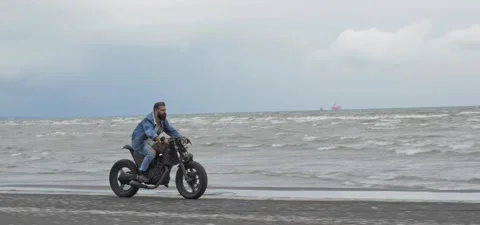 Bearded man in jeans rides old cafe racer motorcycle on sandy beach near sea Stock Footage 140624448